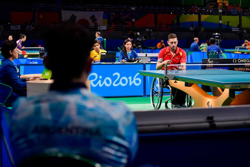 En primer plano, el entrenador de espaldas. En la parte superior derecha, Mauro Depergola a punto de realizar un saque en tenis de mesa en silla de ruedas.