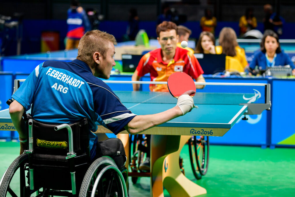 Partido de tenis de mesa en silla de ruedas, donde Fernando Eberhardt, está realizando un saque con la pelota en el aire y su mano atada a la paleta con una venda, frente a su rival coreano.