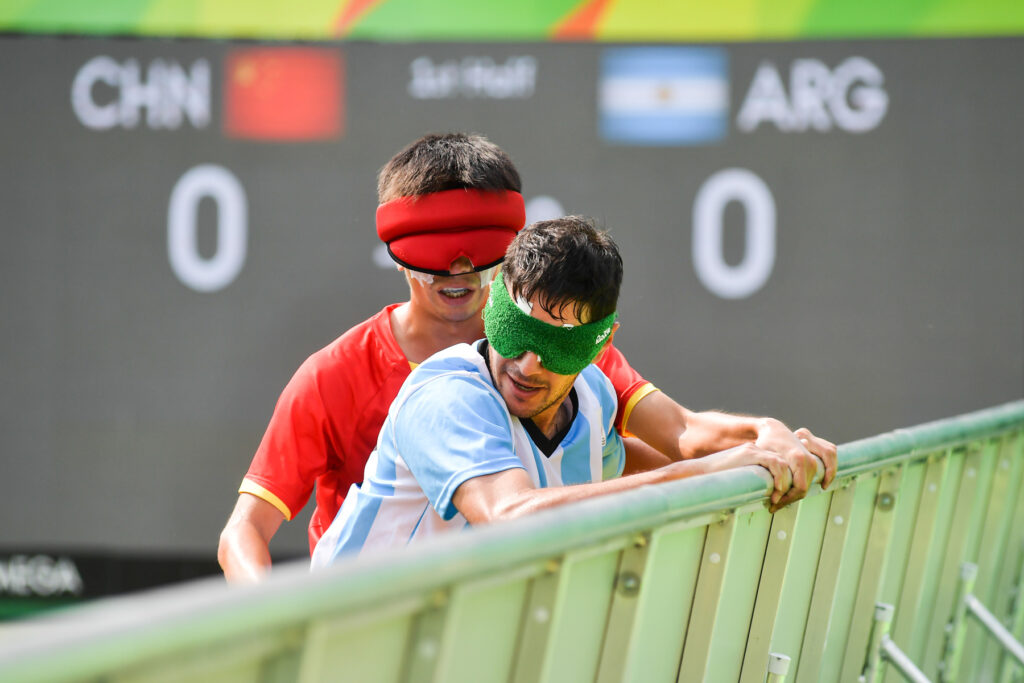 Ángel Deldo, de la Selección Argentina masculina de fútbol para ciegos, lucha contra un jugador chino junto a las vallas del lateral de la cancha. El marcador muestra 0-0.